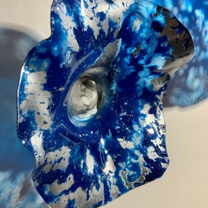 May include: A decorative wall sculpture shaped like a flower with wavy, ruffled edges. The surface features a marbled pattern of deep blue and metallic silver paint, creating a high-contrast, swirling effect across the curved, petal-like form.