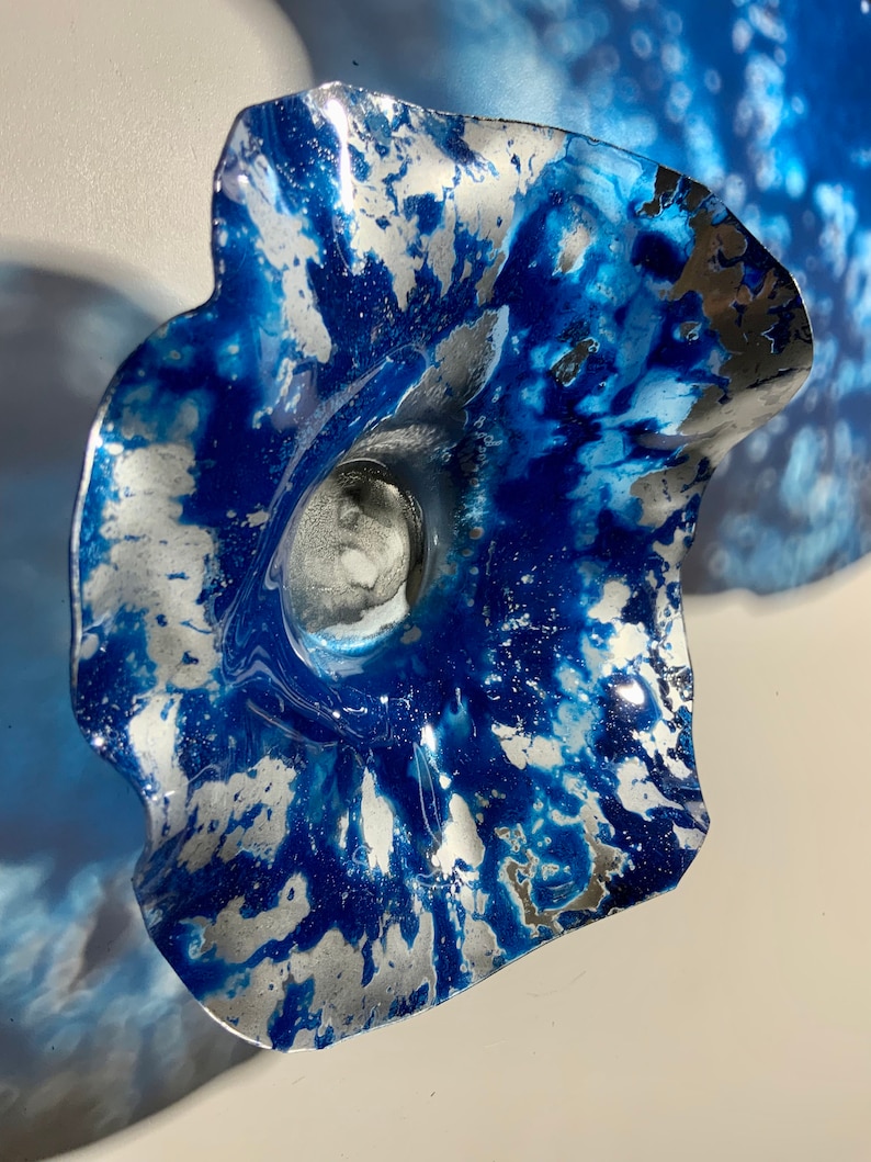 May include: A decorative wall sculpture shaped like a flower with wavy, ruffled edges. The surface features a marbled pattern of deep blue and metallic silver paint, creating a high-contrast, swirling effect across the curved, petal-like form.