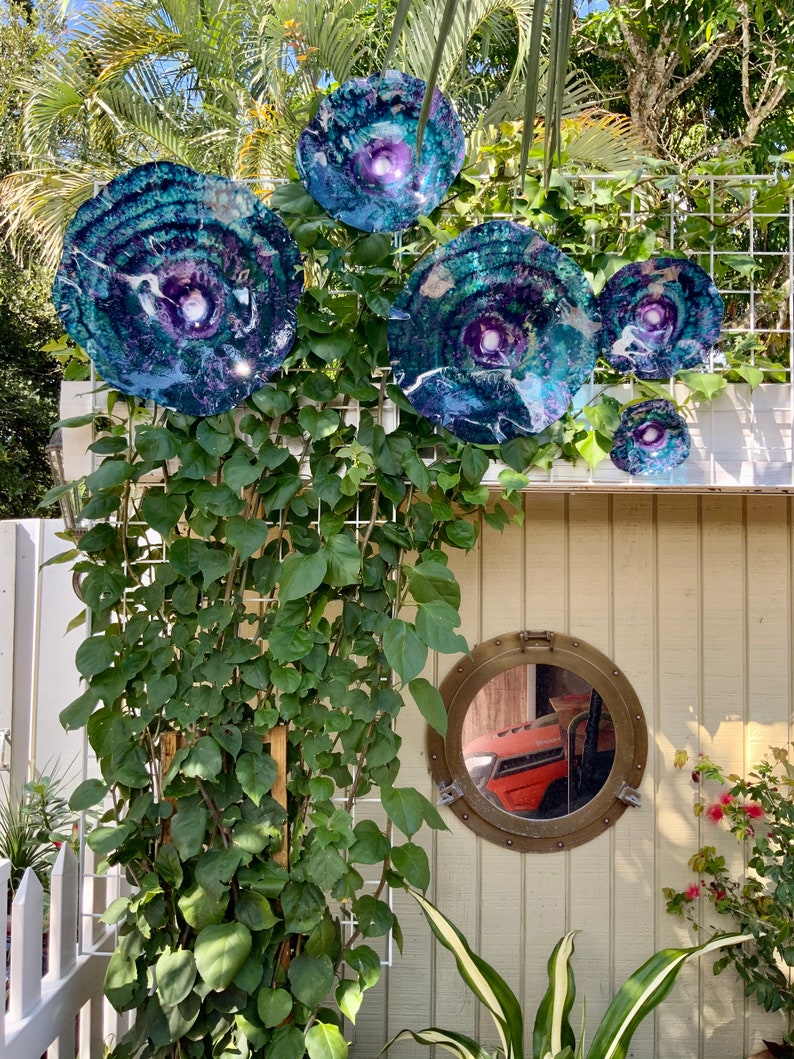 May include: A collection of four circular, multi-coloured glass wall art pieces featuring swirling patterns of deep blue, purple, and teal with white accents, mounted on a white wire trellis above a round, brass-rimmed porthole-style mirror.