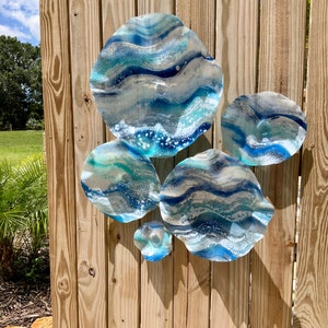 May include: Six abstract wall art pieces with a wave design in shades of blue, white, and silver, mounted on a wooden fence. The pieces evoke ocean waves, creating a coastal theme for home decor.