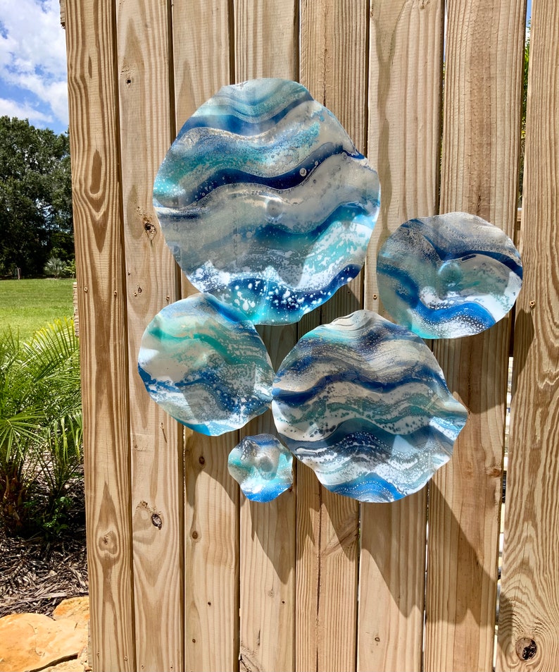 May include: Six abstract wall art pieces with a wave design in shades of blue, white, and silver, mounted on a wooden fence. The pieces evoke ocean waves, creating a coastal theme for home decor.
