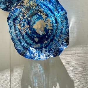 May include: A circular wall-mounted glass sculpture featuring a vibrant blue and white marbled pattern with metallic gold accents. The piece has a ruffled, wavy edge and a translucent center, creating a dynamic, layered appearance with light.