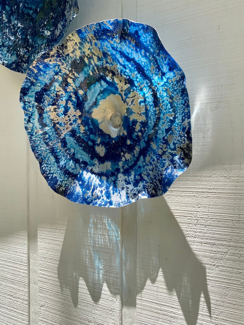 May include: A circular wall-mounted glass sculpture featuring a vibrant blue and white marbled pattern with metallic gold accents. The piece has a ruffled, wavy edge and a translucent center, creating a dynamic, layered appearance with light.