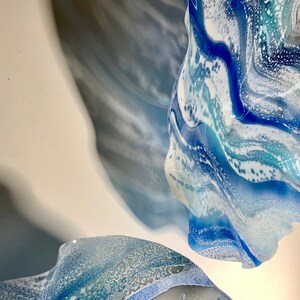 May include: Abstract artwork showing two flowing, organic shapes in shades of blue and white. The forms appear to be made of glass or a similar translucent material, with a marbled effect. The artwork is mounted on a white surface.
