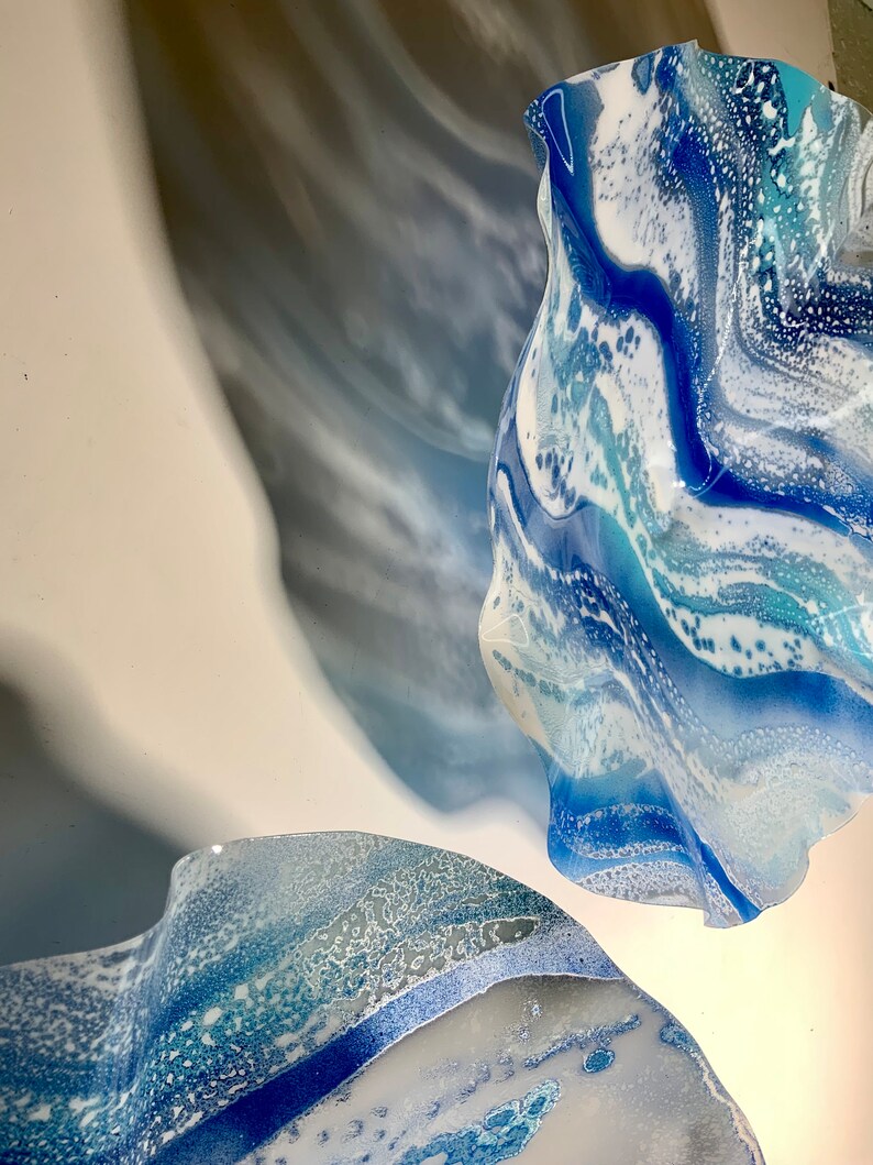 May include: Abstract artwork showing two flowing, organic shapes in shades of blue and white. The forms appear to be made of glass or a similar translucent material, with a marbled effect. The artwork is mounted on a white surface.