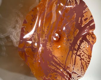 Könnte beinhalten: Ein kreisförmiges Wandkunstobjekt aus Glas mit einem wellenförmigen, organischen Rand. Die Oberfläche zeigt ein marmoriertes Muster in tiefen Mauvetönen und leuchtendem Orange mit gesprenkelten Details und einem zentralen Knopf aus Klarglas.
