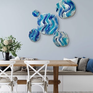 May include: A wall art installation featuring five abstract, round and square pieces with a blue, white, and turquoise wave pattern. The art is mounted on a light blue wall above a wooden table and white chairs.