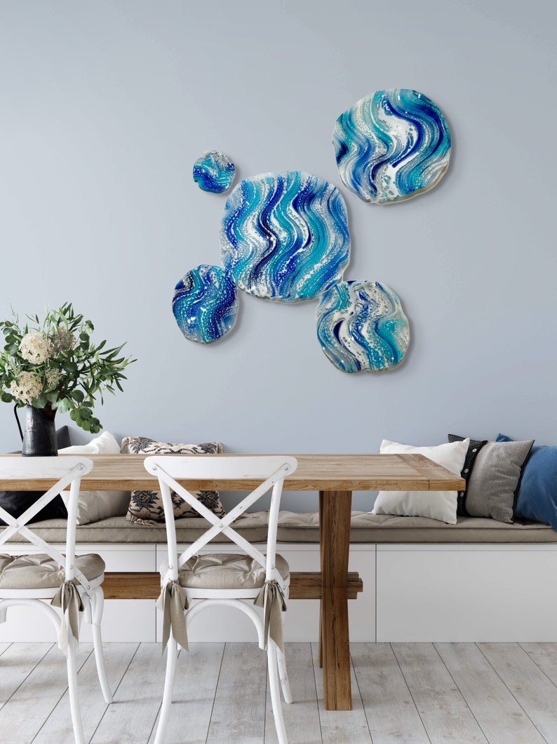 May include: A wall art installation featuring five abstract, round and square pieces with a blue, white, and turquoise wave pattern. The art is mounted on a light blue wall above a wooden table and white chairs.