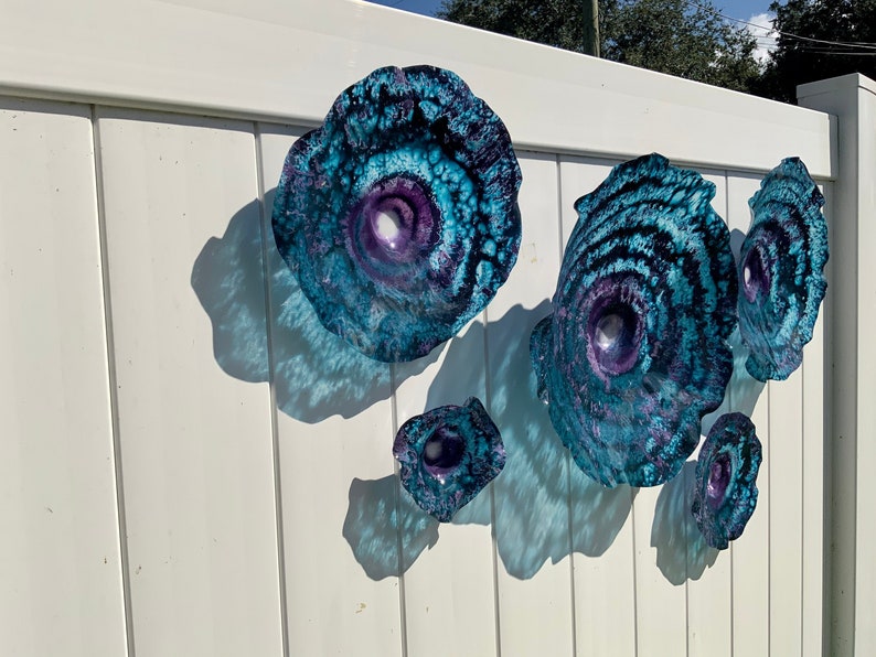 May include: A collection of five circular wall art pieces featuring a swirling pattern of deep blue, turquoise, and purple hues. Each piece has a concave centre and a textured, organic edge, creating a layered appearance with varying sizes.