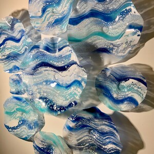 May include: A collection of decorative glass wall art pieces in various sizes and shapes. The pieces feature a swirling design in shades of blue and white, resembling ocean waves. The art is mounted on a white wall, creating a striking visual display.