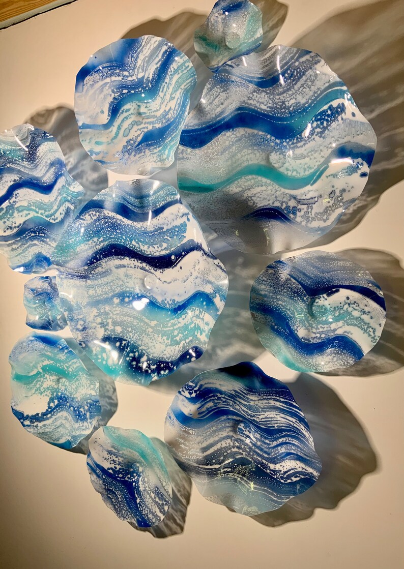 May include: A collection of decorative glass wall art pieces in various sizes and shapes. The pieces feature a swirling design in shades of blue and white, resembling ocean waves. The art is mounted on a white wall, creating a striking visual display.