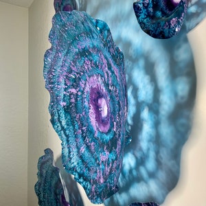 May include: A collection of wall-mounted circular art pieces featuring a vibrant colour palette of deep purple, bright teal, and dark blue. The surfaces display a swirling, marbled pattern with textured edges, creating a layered effect on the wall.