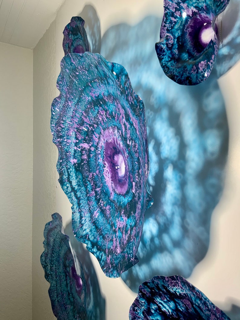 May include: A collection of wall-mounted circular art pieces featuring a vibrant colour palette of deep purple, bright teal, and dark blue. The surfaces display a swirling, marbled pattern with textured edges, creating a layered effect on the wall.