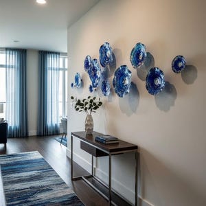 May include: A collection of eleven circular blue glass wall sculptures with textured, rippled surfaces arranged in a cluster on a plain wall above a dark wood console table that holds a vase with green foliage and a stack of books.