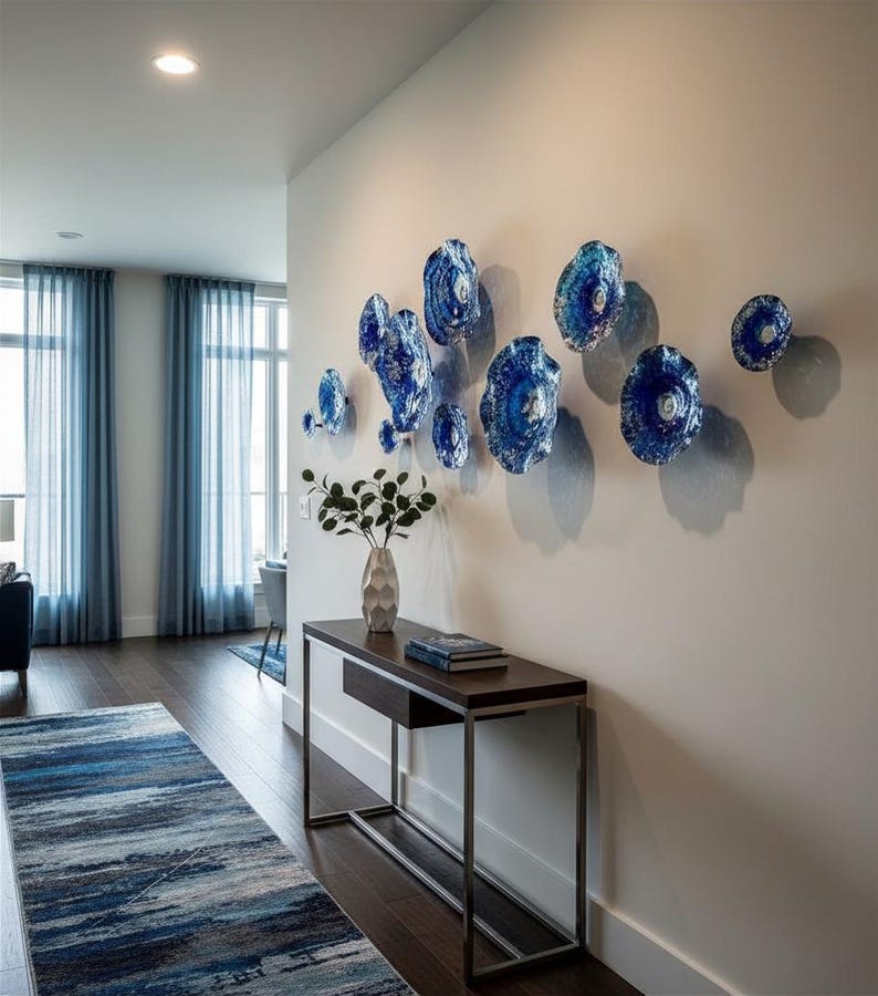 May include: A collection of eleven circular blue glass wall sculptures with textured, rippled surfaces arranged in a cluster on a plain wall above a dark wood console table that holds a vase with green foliage and a stack of books.