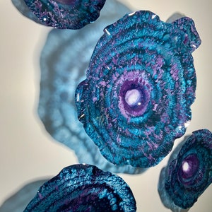 May include: Four circular resin coasters featuring a swirling pattern of deep blue, teal, and purple hues. Each piece has a prominent white centre surrounded by concentric rings of colour, creating a textured, agate-like appearance with a glossy finish.