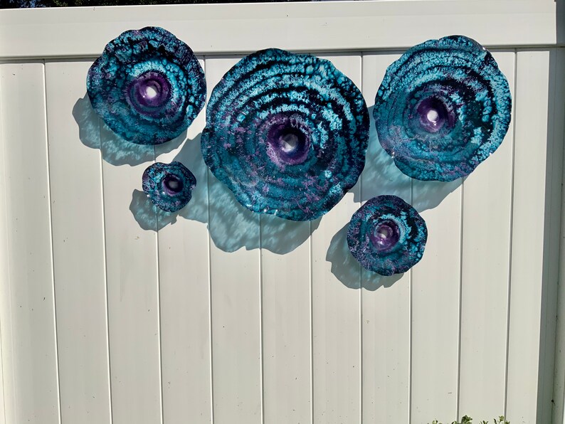 May include: A set of five circular wall art pieces featuring a textured, swirling pattern in shades of deep blue, teal, and purple. Each piece has a concave centre and a translucent, glass-like appearance with organic, irregular edges.