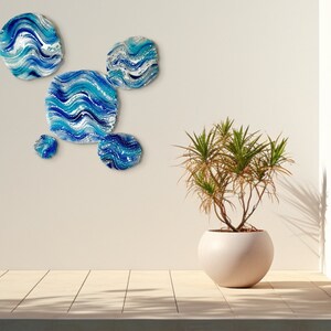 May include: A collection of five circular wall art pieces with a blue and white wave design. The artwork is displayed on a neutral wall. A potted plant in a spherical pot is also visible. The plant is in a pot with a diameter of approximately 40 cm.