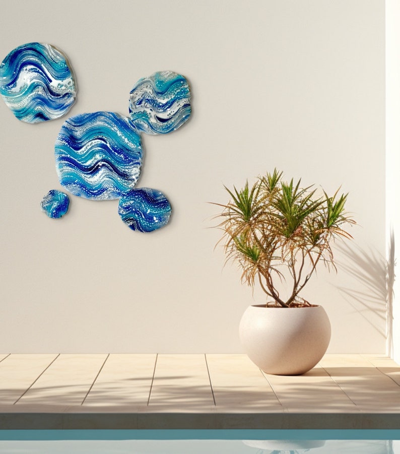 May include: A collection of five circular wall art pieces with a blue and white wave design. The artwork is displayed on a neutral wall. A potted plant in a spherical pot is also visible. The plant is in a pot with a diameter of approximately 40 cm.