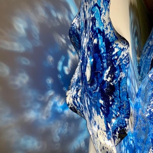 May include: A wall-mounted glass sculpture featuring a wavy, organic shape with a vibrant blue and white marbled pattern. The surface has a glossy finish that reflects light, creating intricate blue shadows on the adjacent wall surface.