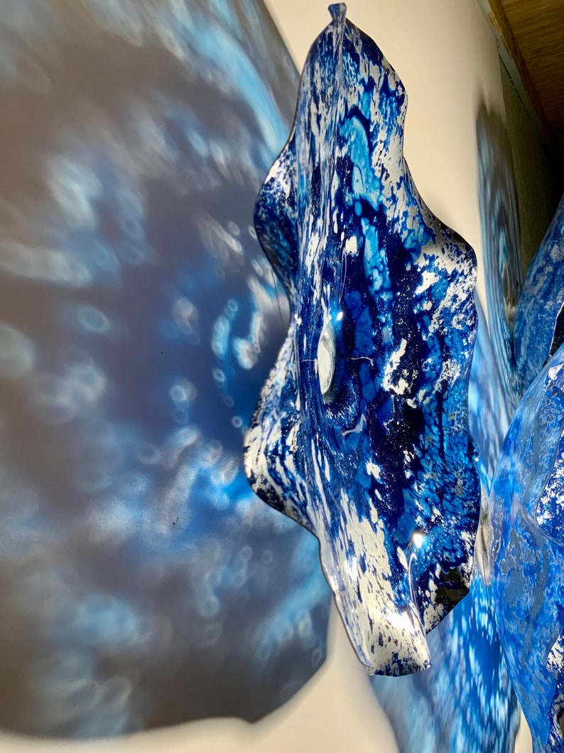 May include: A wall-mounted glass sculpture featuring a wavy, organic shape with a vibrant blue and white marbled pattern. The surface has a glossy finish that reflects light, creating intricate blue shadows on the adjacent wall surface.