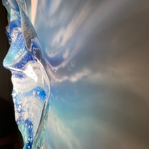 May include: A wall-mounted art piece with a layered, flowing design. The artwork features shades of blue and white, with a textured, wave-like appearance. The light creates shadows on the wall, enhancing the piece's depth and form.