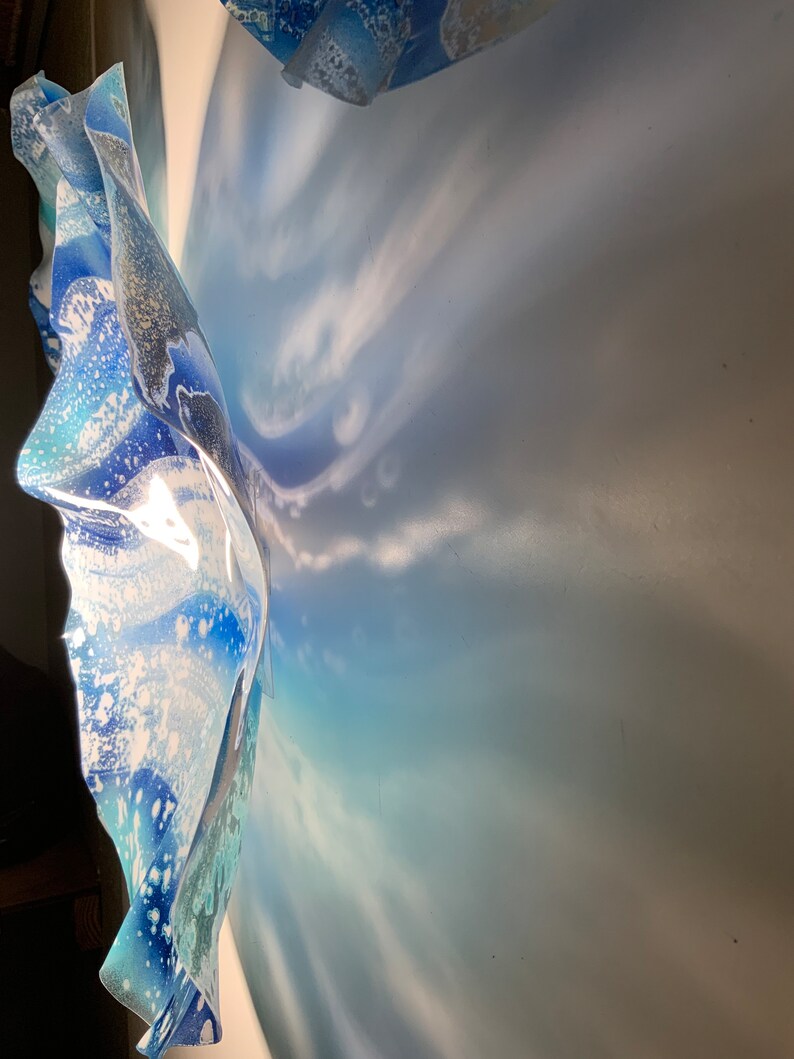 May include: A wall-mounted art piece with a layered, flowing design. The artwork features shades of blue and white, with a textured, wave-like appearance. The light creates shadows on the wall, enhancing the piece's depth and form.