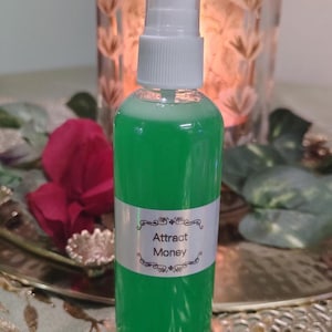 May include: A clear spray bottle filled with bright green liquid. The bottle has a white label with the words "Attract Money" in elegant script. The bottle is topped with a white spray nozzle.
