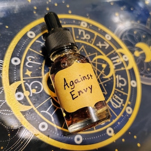 May include: A small glass bottle with a black dropper top and a brown liquid inside. A tan label on the bottle reads "Against Envy" in black ink. The bottle sits on a dark blue surface with a gold astrological design.