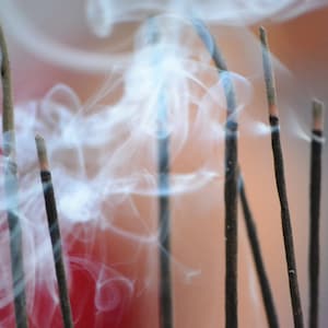 May include: Close-up of several incense sticks burning, with wisps of white smoke curling upwards. The sticks are dark brown with a red tip, set against a blurred background of red and peach tones.