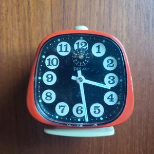Könnte beinhalten: Roter Wecker mit schwarzem Zifferblatt und weißen Zahlen. Die Uhr hat einen kleinen Sekundenzeiger und die Worte "Peter" und "Made in Germany" sind auf dem Zifferblatt aufgedruckt.