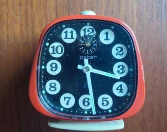 May include: Red alarm clock with black face and white numbers. The clock has a small second hand and the words "Peter" and "Made in Germany" are printed on the face.