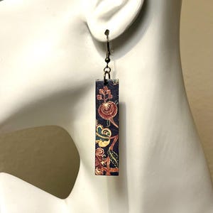 May include: A single rectangular earring with a dark blue background and a floral pattern in red, yellow, and green. The earring is suspended from a dark metal hook. The earring is displayed on a white mannequin head.