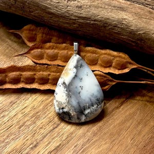 May include: A teardrop-shaped pendant with a silver bail. The pendant features a white, gray, and black patterned stone. The word "Riversidebliss" is written in cursive at the bottom of the image. The pendant is displayed on a wooden surface.