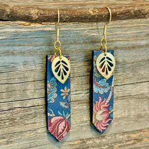 May include: A pair of dangle earrings with a floral pattern. The earrings feature a gold-colored leaf charm and a rectangular pendant with a blue background and a pink and white floral design. The earrings are suspended from gold-colored hooks.