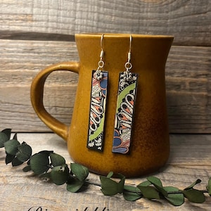 May include: A pair of rectangular dangle earrings with intricate floral and paisley patterns in black, green, orange, and blue. The earrings hang from silver hooks and are displayed against a brown mug. The text "Riversidebliss" is at the bottom.