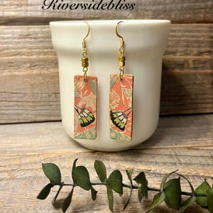 May include: A pair of rectangular earrings with a floral and butterfly design. The earrings are suspended from gold-colored hooks and feature a mix of coral, green, and white colors. The text "Riversidebliss" is visible at the top.