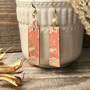 May include: A pair of rectangular earrings with a floral design. The earrings have a salmon-colored background with gold floral patterns. They hang from gold-colored hooks and have a small, round, cream-colored bead. The earrings are displayed in front of a white ceramic pot.