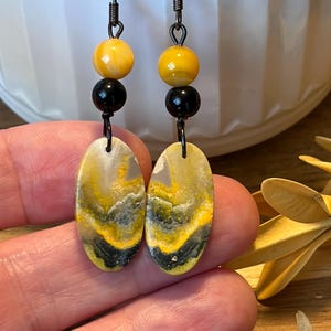 May include: Dangle earrings featuring oval-shaped bumblebee jasper stones with yellow, gray, and black patterns. Each earring includes a small black bead and a yellow bead suspended from a dark metal hook, creating a layered, multi-textured appearance.