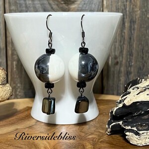 May include: Black and white ceramic sphere earrings with black beads and a square-shaped dangle. The earrings are hanging from a white bowl.