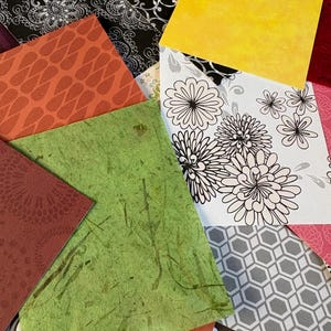 May include: A collection of overlapping square paper sheets featuring diverse patterns and textures, including a bright yellow sheet, a green fibrous paper, a white sheet with black floral line art, and various geometric and damask prints.