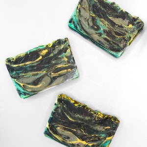 May include: Three rectangular bars of soap featuring a marbled pattern of black, deep teal, and metallic gold swirls. Each bar has a smooth, flat surface with a slightly textured, uneven top edge, showcasing a complex blend of dark and vibrant colors.