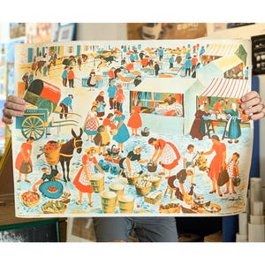 May include: A vintage print showing a busy market scene. The artwork features many figures in colourful clothing, stalls with striped awnings, and a donkey carrying baskets. The overall colour palette includes reds, blues, and yellows.