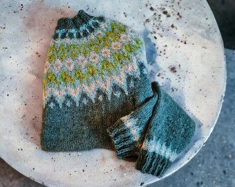 May include: A green, blue, and pink knitted sweater with a geometric pattern. The sweater is folded and placed on a light gray surface.