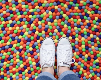May include: A pair of white canvas shoes with laces are standing on a colourful round rug made of felt balls. The rug is a mix of red, orange, yellow, green, blue, and purple felt balls.