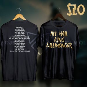 Puede incluir: Camisetas negras con texto dorado. Una camiseta dice "ALL HAIL KING KILLMONGER". La otra camiseta tiene una lista de frases. Ambas camisetas están colgadas en perchas de madera. El precio es de 20 €.