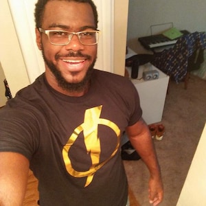 May include: A man wearing a black t-shirt with a gold lightning bolt symbol. He is wearing glasses and has a beard.