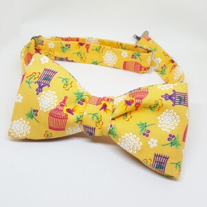 May include: A yellow bow tie with a repeating pattern of white flowers, birds in cages, and green leaves.
