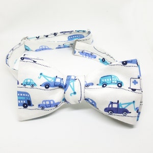 May include: White bow tie with a blue and white pattern of cars, trucks, and buses. The bow tie is made of fabric and has a white adjustable strap.