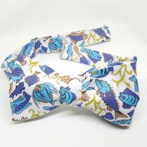 May include: A white bow tie with a blue and green fish pattern. The bow tie is made of fabric and has a classic design.